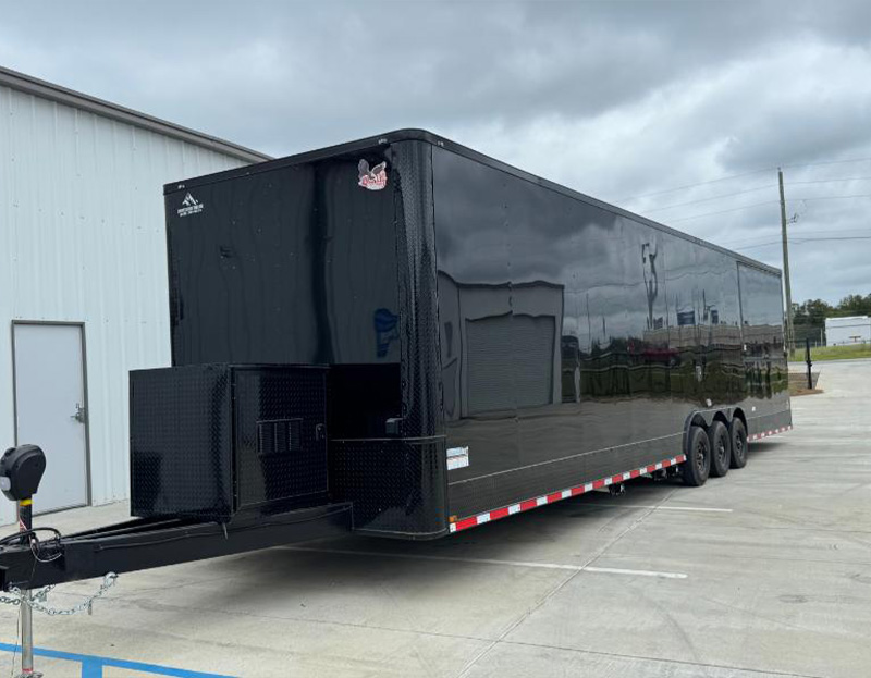 The image shows a large black enclosed trailer parked on an asphalt surface next to a building with a grey roof.