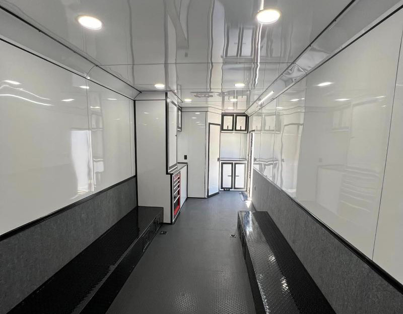 The image shows the interior of a professional racing car transporter with a white and black color scheme, featuring a large storage area, a workbench, and various equipment, including tools and a red fire extinguisher.
